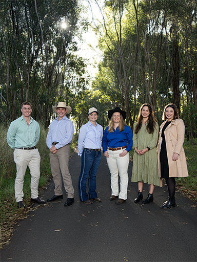 Six young finalists of the RAS Foundation Rural Scholarhip standing outside on a tree-lined path, smiling and dressed in smart rural attire.