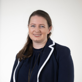 Headshot of Goat Committee Chair Sarah Hanlon, smiling and wearing a navy jacket with white trim over a dark top.