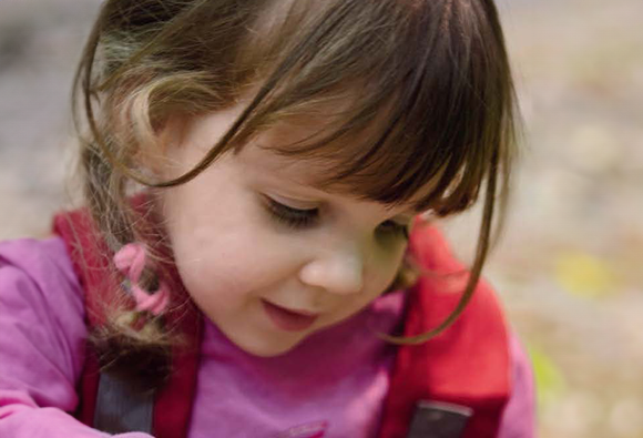 A young girl in a pink jumper looking down with a gentle smile outdoors.