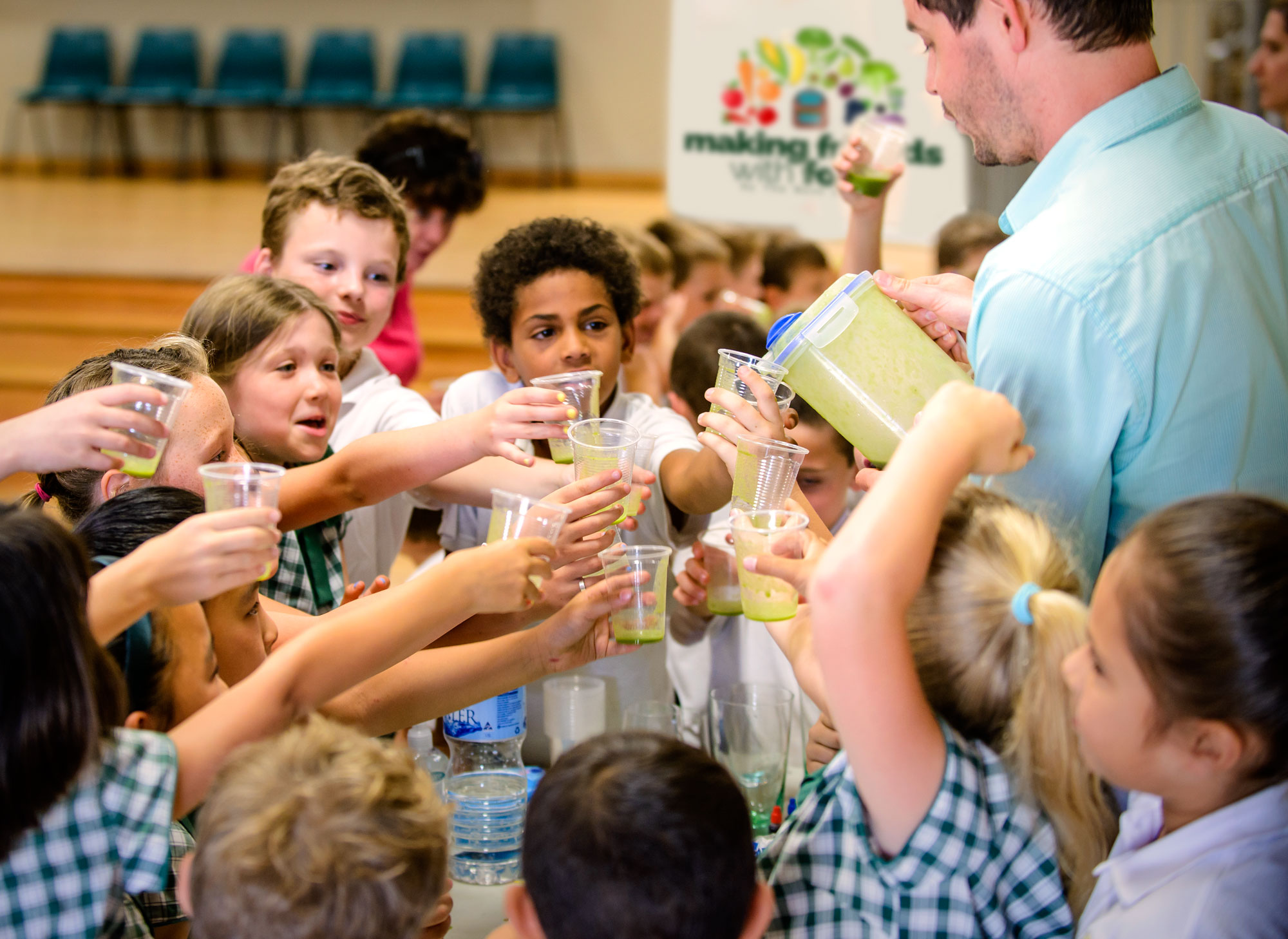 School children in uniform raising cups of green smoothies while an adult pours from a jug.