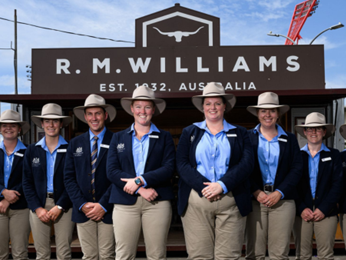 Five RAS Rural Achiever finalists in matching blue shirts and beige trousers stand smiling outside an R.M. Williams building.