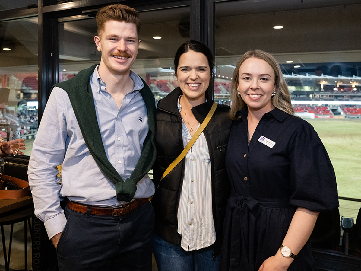 Three smiling people stand indoors near a stadium window during Hats Off to the Royal.