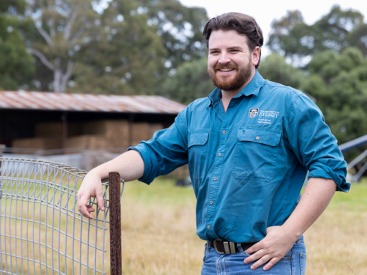 RAS Youth Group member Jack Mayhew leans on a wire fence, smiling in a rural paddock with a hay shed behind him.