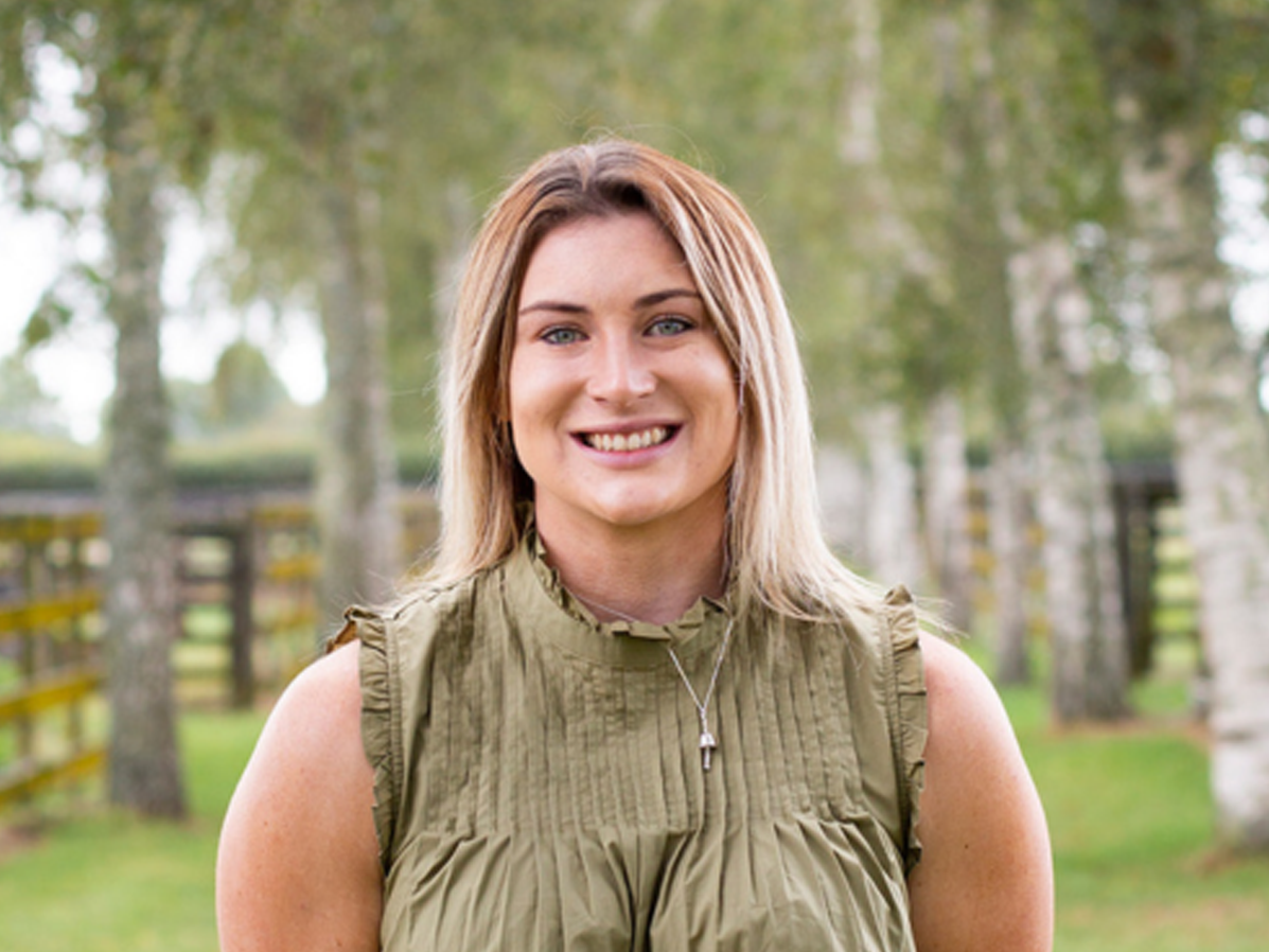 RAS Youth Group member Madison Tims with light brown hair, wearing a sleeveless green blouse smiling with trees in the background.