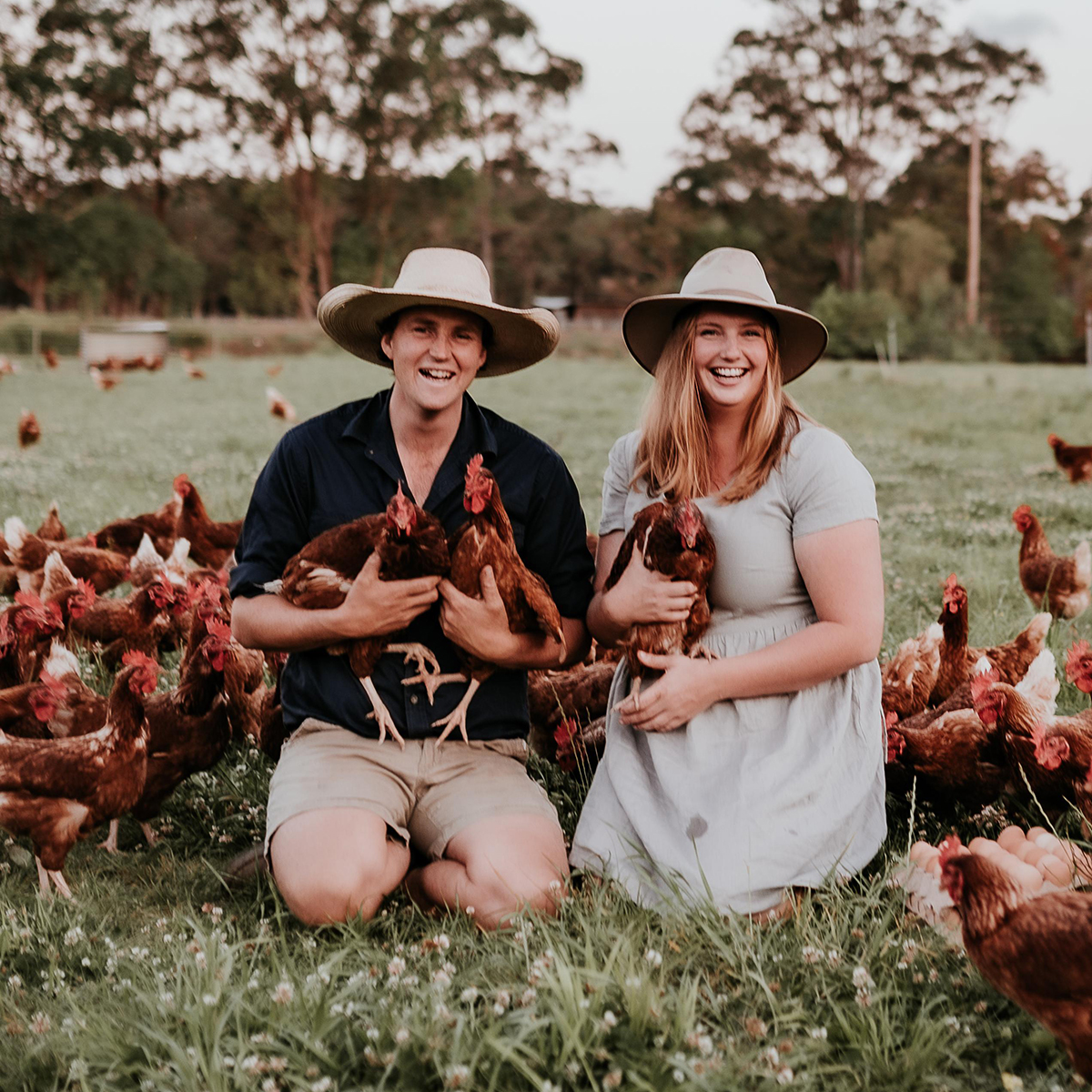 Two people kneeling in a paddock surrounded by chickens, each holding hens and smiling at the camera.